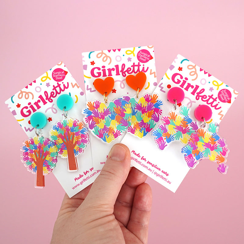 A hand holding 3 pairs of Harmony Day teacher acrylic dangle earrings with rainbow hands