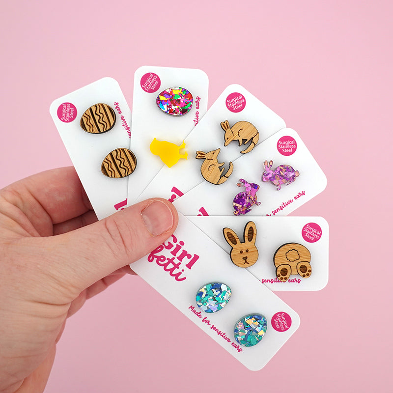 A hand holding 6 pairs of acrylic Easter stud earrings including Easter eggs, a chicken, a bilby and Easter bunny rabbits