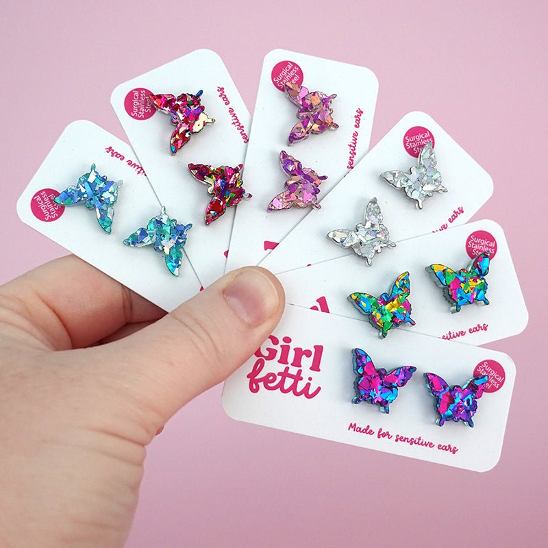 A hand holding 6 pairs of glitter acrylic butterfly stud earrings