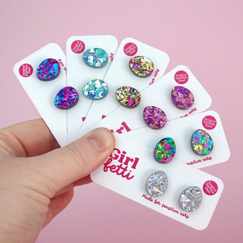 A hand holding 6 pairs of glitter acrylic Easter egg stud earrings