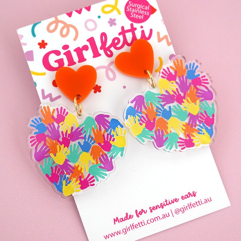 A pair of acrylic teacher dangle earrings for Harmony Day with rainbow hands printed on them in the shape of a heart